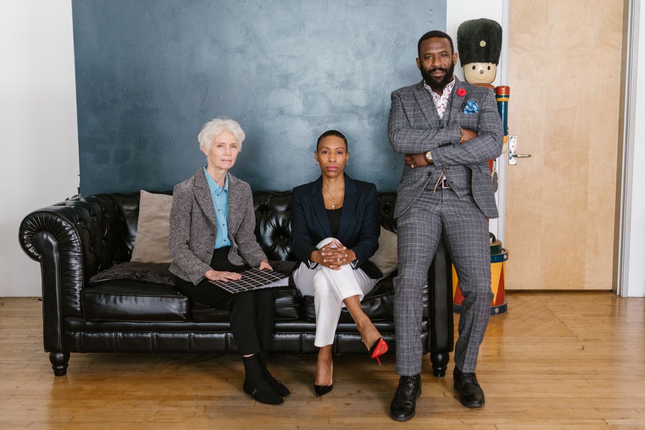 who-we-are Three diverse professionals posing confidently on a black sofa in a stylish office setting.