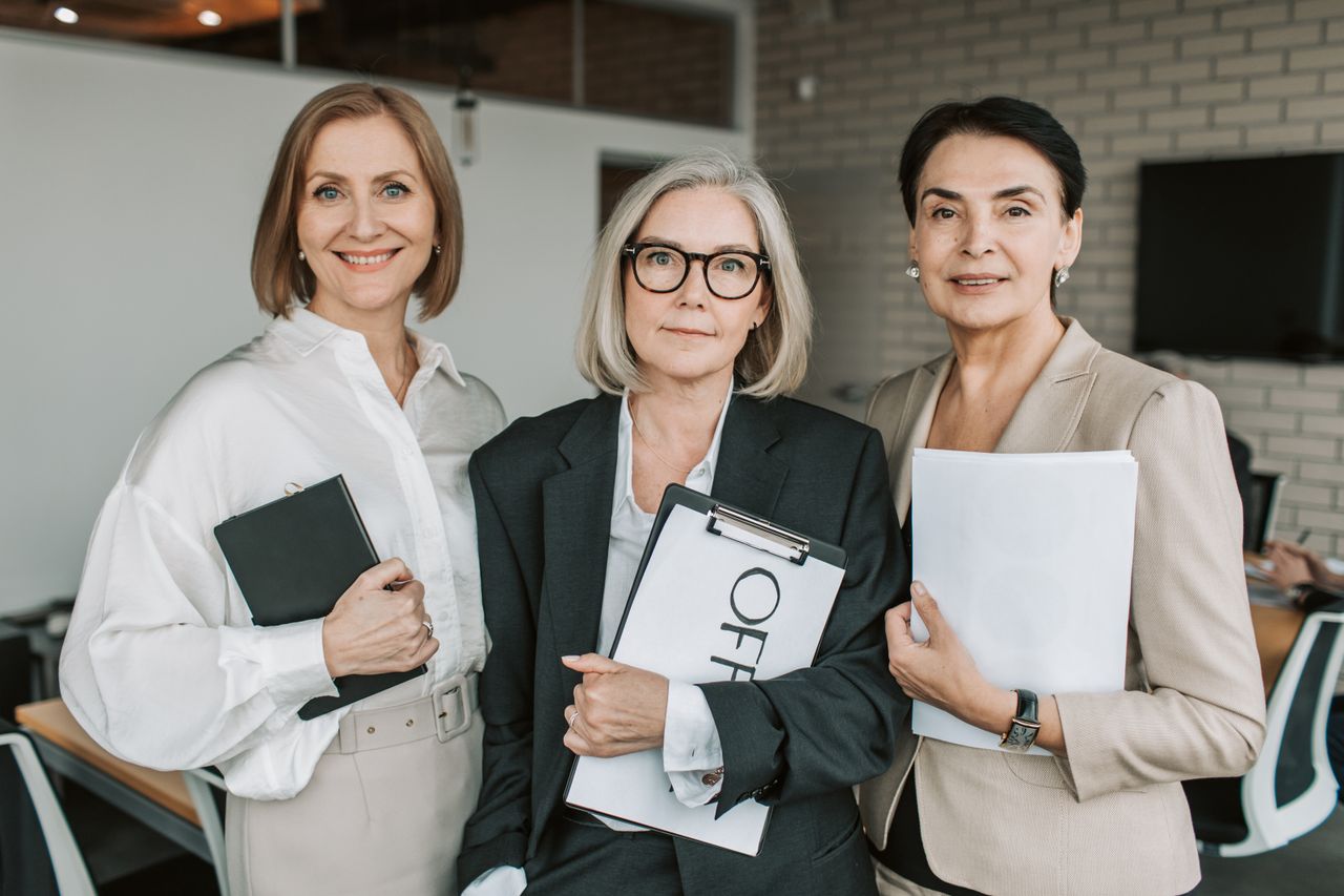 services-02 Three confident businesswomen in professional attire holding papers in an office setting.