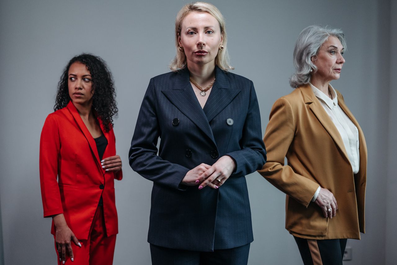services-01 Three businesswomen in suits pose confidently on a neutral background, showcasing diversity.